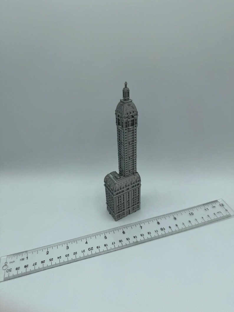 May include: A miniature model of a tall, gray building with a pointed roof and many windows. The model is made of metal and is sitting on a white surface next to a ruler.