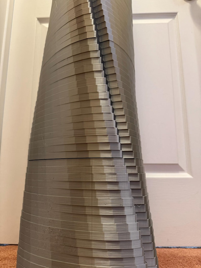 May include: A 3D printed sculpture made of stacked layers of plastic, creating a unique and abstract design. The sculpture is a light grey colour and has a smooth, slightly textured surface.