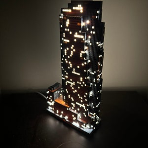May include: A detailed model of a skyscraper with illuminated windows, made of wood and plastic. The model is lit from within, creating a warm glow.