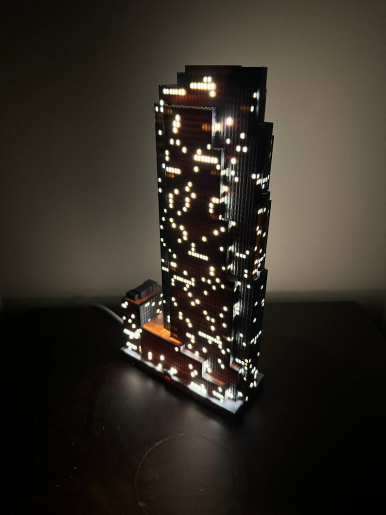 May include: A detailed model of a skyscraper with illuminated windows, made of wood and plastic. The model is lit from within, creating a warm glow.