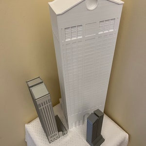 May include: Three white 3D printed model buildings. The tallest building is white and has a rounded roof. The two smaller buildings are gray and have flat roofs.