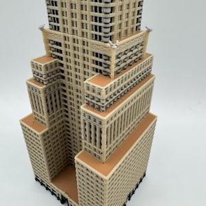 May include: A beige and brown architectural model of a skyscraper. The model has a tiered design with multiple rectangular sections. The windows are represented by dark, rectangular openings. The building has a detailed, textured facade.