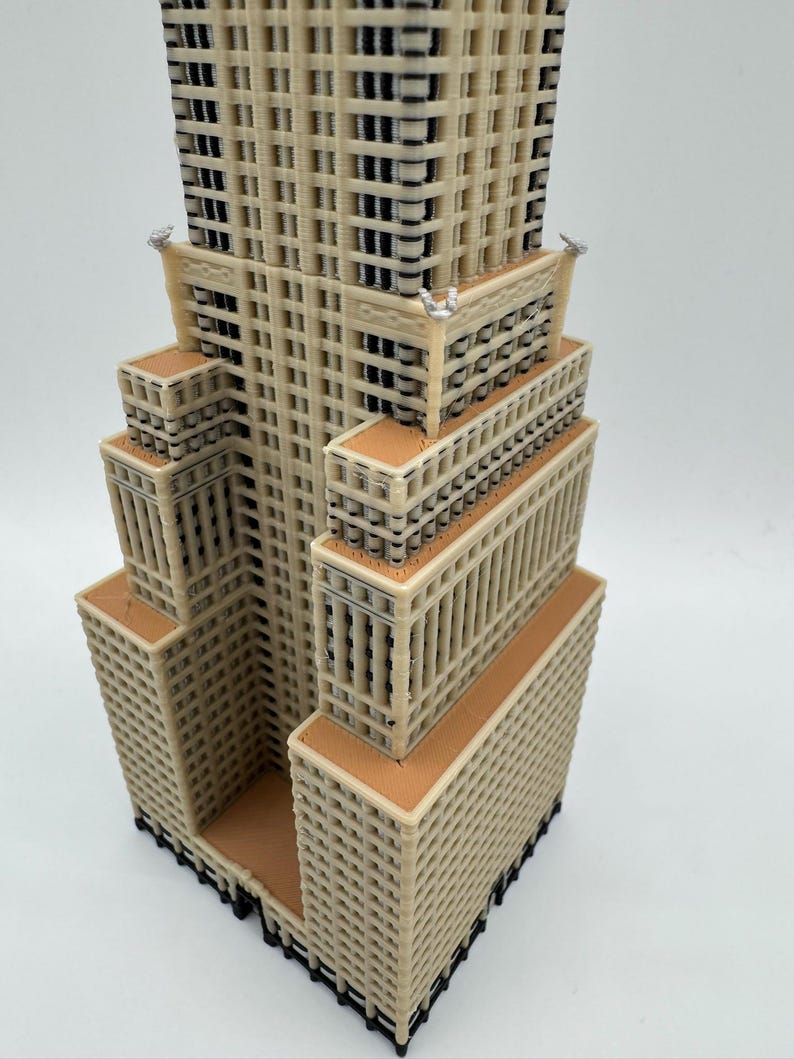 May include: A beige and brown architectural model of a skyscraper. The model has a tiered design with multiple rectangular sections. The windows are represented by dark, rectangular openings. The building has a detailed, textured facade.