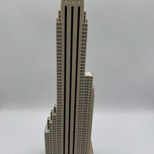 May include: A beige skyscraper model with black vertical stripes and numerous rectangular windows. The building has a stepped design and a flat roof. The model is a miniature representation of a famous building.