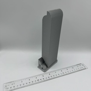 May include: A gray 3D-printed model of a skyscraper with a grid-like facade. The building is next to a clear ruler, approximately 12 inches long, for scale. The model has a curved top and a detailed base.