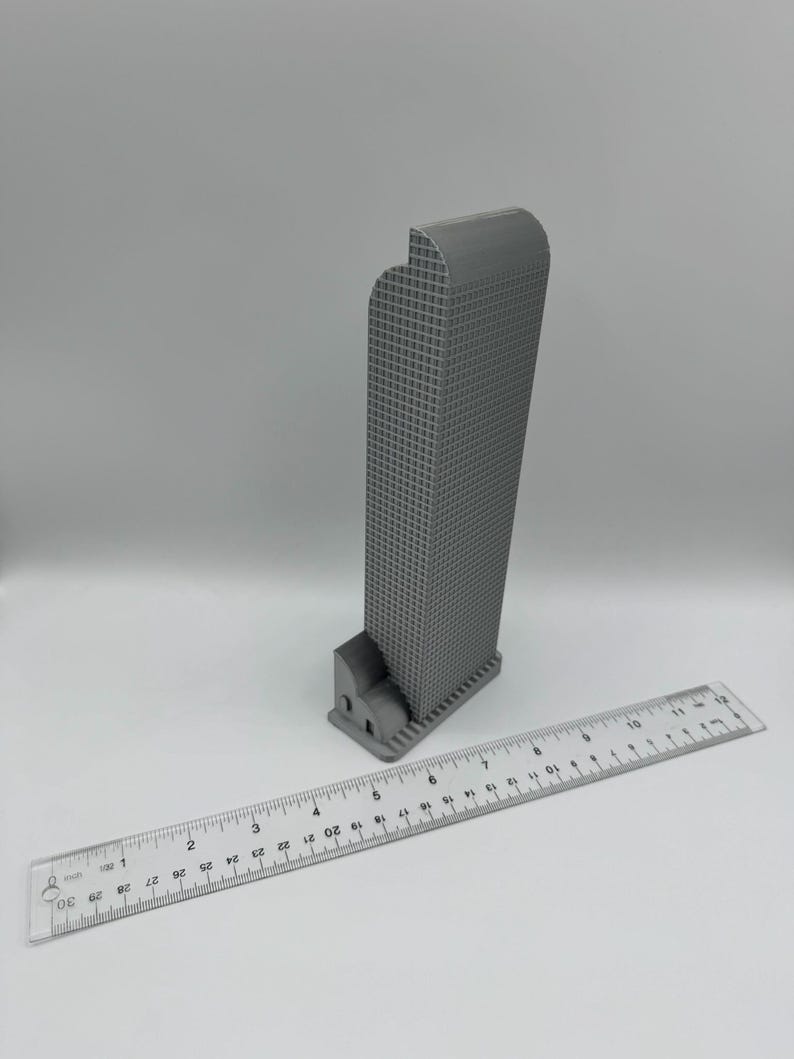 May include: A gray 3D-printed model of a skyscraper with a grid-like facade. The building is next to a clear ruler, approximately 12 inches long, for scale. The model has a curved top and a detailed base.