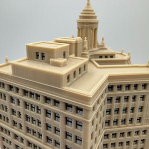 Op de afbeelding: Een gedetailleerd 3D-geprint model van een hoog gebouw met een decoratief dak en vele ramen. Het gebouw is gemaakt van lichtbruin plastic en lijkt een replica te zijn van een historische wolkenkrabber.
