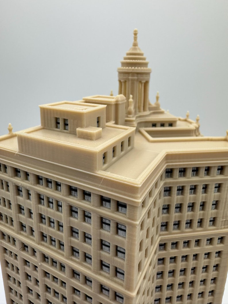 Op de afbeelding: Een gedetailleerd 3D-geprint model van een hoog gebouw met een decoratief dak en vele ramen. Het gebouw is gemaakt van lichtbruin plastic en lijkt een replica te zijn van een historische wolkenkrabber.