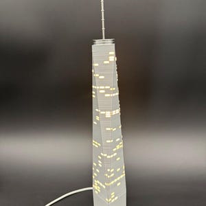 May include: A gray miniature replica of a skyscraper with illuminated windows. The model has a long antenna and a white cord. The building is a detailed representation of a modern architectural design.