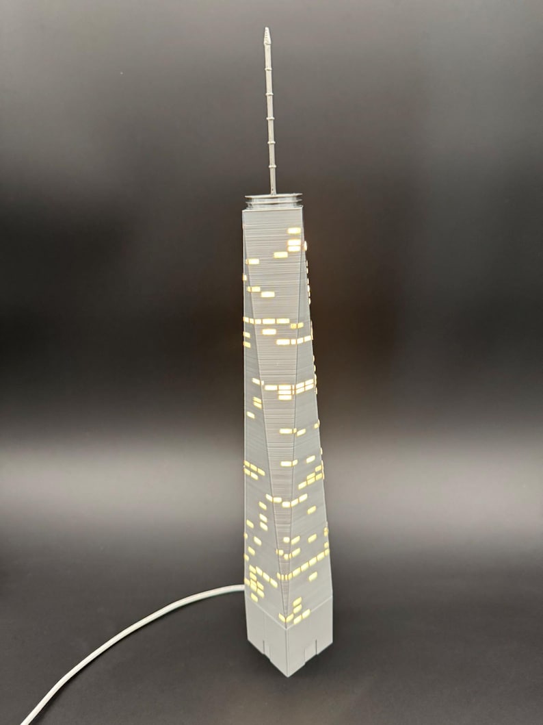 May include: A gray miniature replica of a skyscraper with illuminated windows. The model has a long antenna and a white cord. The building is a detailed representation of a modern architectural design.