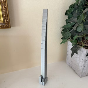 May include: A silver-coloured architectural model, rectangular in shape, with a series of stacked sections. The model is displayed on a white surface. A decorative gold-framed picture and a potted plant are in the background.