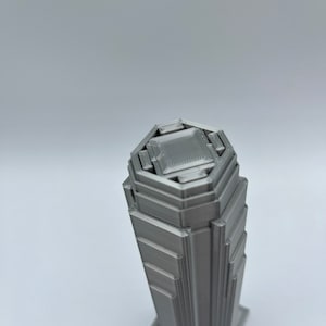 May include: A 3D printed model of a skyscraper with a hexagonal base and a layered design. The model is made of a silver-colored material.