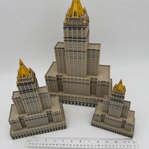 May include: Three-dimensional model of a building with a beige facade and gold-colored spire. The model is placed on a white surface with a ruler for scale. The building has many windows and architectural details.