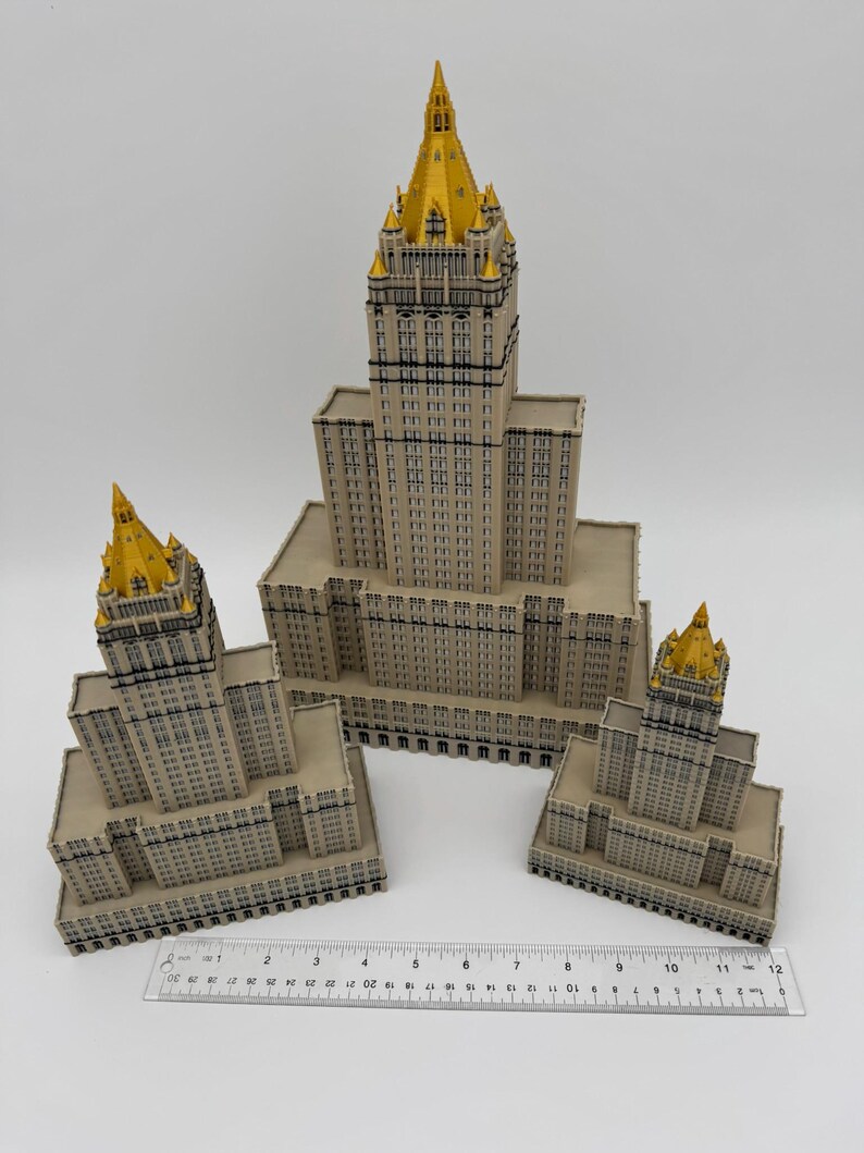 May include: Three-dimensional model of a building with a beige facade and gold-colored spire. The model is placed on a white surface with a ruler for scale. The building has many windows and architectural details.