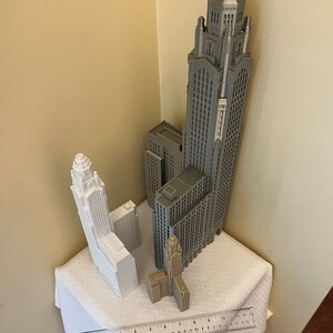 May include: Three 3D printed model buildings, one gray and two white, on a white tablecloth with a ruler. The tallest building is gray and has a pointed spire. The other two buildings are shorter and have flat roofs.