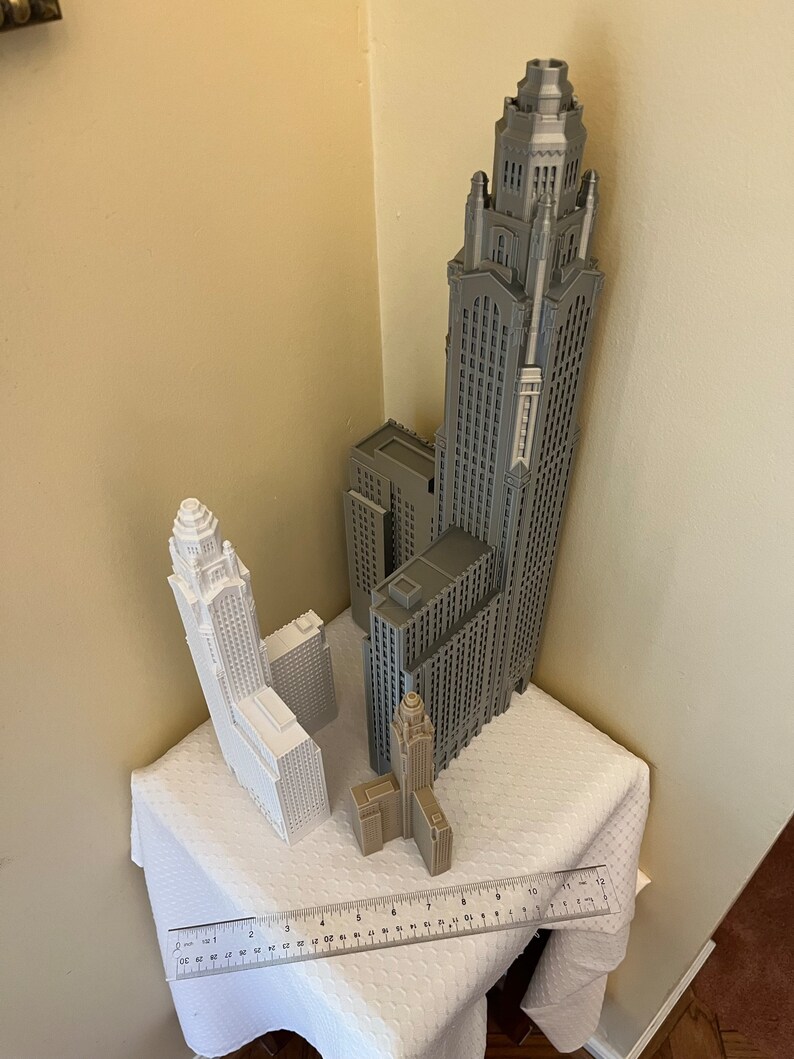 May include: Three 3D printed model buildings, one gray and two white, on a white tablecloth with a ruler. The tallest building is gray and has a pointed spire. The other two buildings are shorter and have flat roofs.