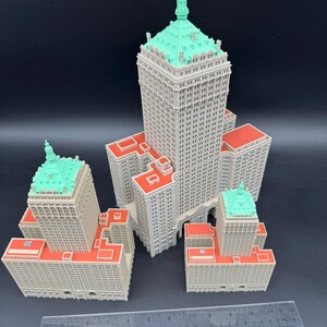 May include: A collection of miniature building models. The buildings are light gray with red rooftops and green accents. A ruler is placed in front of the models for scale.