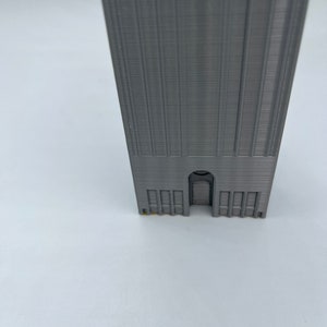 May include: A gray 3D printed model of a tall building with a single arched doorway and a row of windows at the base.
