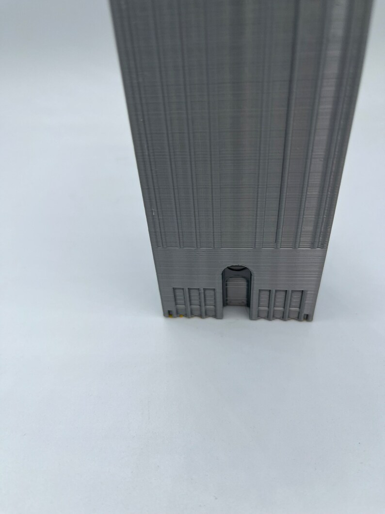 May include: A gray 3D printed model of a tall building with a single arched doorway and a row of windows at the base.