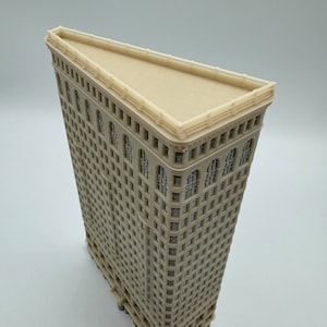 May include: A beige model of the Flatiron Building in New York City. The model is made of plastic and has a detailed facade with windows and decorative elements.