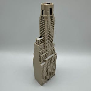 May include: A beige 3D printed architectural model of a skyscraper featuring a stepped design with multiple levels, a grid of small square windows across the facade, and a central tower section that rises above the lower tiered building blocks.