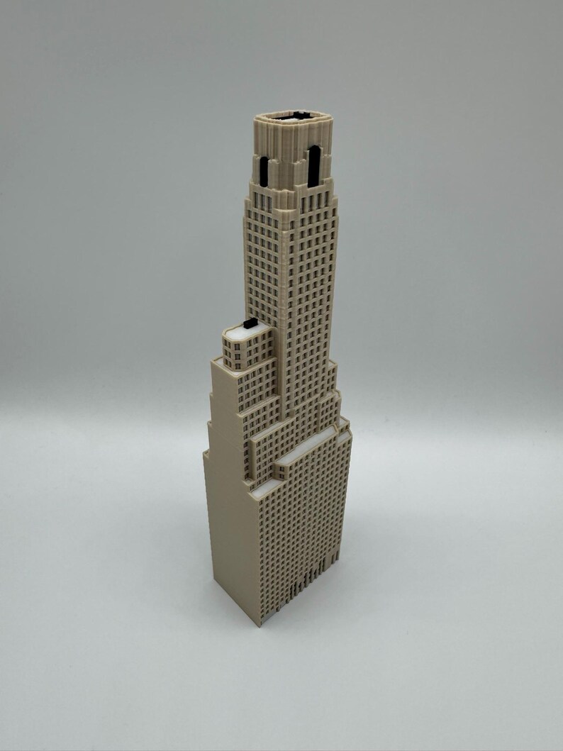 May include: A beige 3D printed architectural model of a skyscraper featuring a stepped design with multiple levels, a grid of small square windows across the facade, and a central tower section that rises above the lower tiered building blocks.