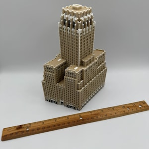 May include: A tan-coloured miniature building with a detailed architectural design. The model has a tall central tower and multiple lower sections. A wooden ruler is placed next to the building, measuring up to 30 cm.