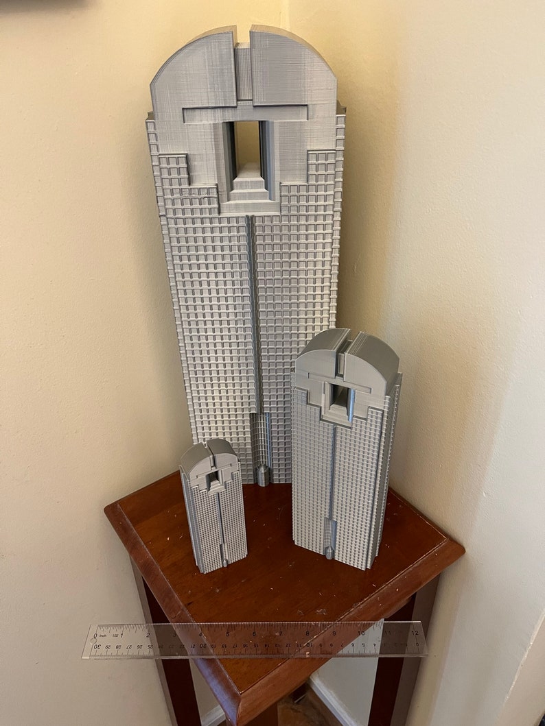 May include: Three grey 3D printed models of a skyscraper. The models are different sizes and are arranged on a wooden table.