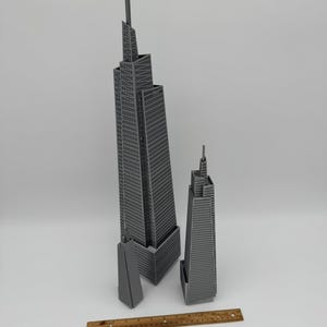 May include: Three gray miniature skyscraper models of varying sizes. The tallest model has a spire and is approximately 18 inches tall. A wooden ruler is placed at the bottom of the image for scale.