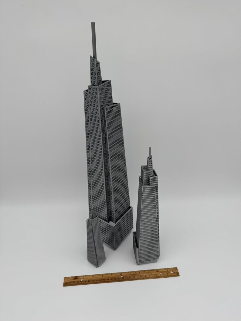 May include: Three gray miniature skyscraper models of varying sizes. The tallest model has a spire and is approximately 18 inches tall. A wooden ruler is placed at the bottom of the image for scale.