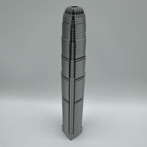 May include: A silver-colored, miniature skyscraper model with a detailed, textured facade. The building has a rectangular base and a tiered design, with a textured top. The model is a representation of a famous building.