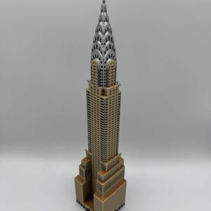 May include: A detailed miniature of the Chrysler Building, showcasing its iconic Art Deco design. The model features a tan facade, silver spire, and intricate architectural details. A collectible item, perfect for display.