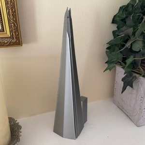 May include: A silver model of the Shard, a skyscraper in London, England. The model is made of metal and has a sharp, pointed top.