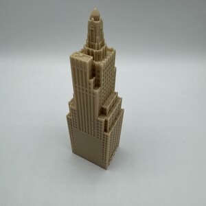 May include: A tan-colored miniature skyscraper model. The building has a detailed facade with multiple levels, windows, and architectural features. The top of the building has a decorative spire.
