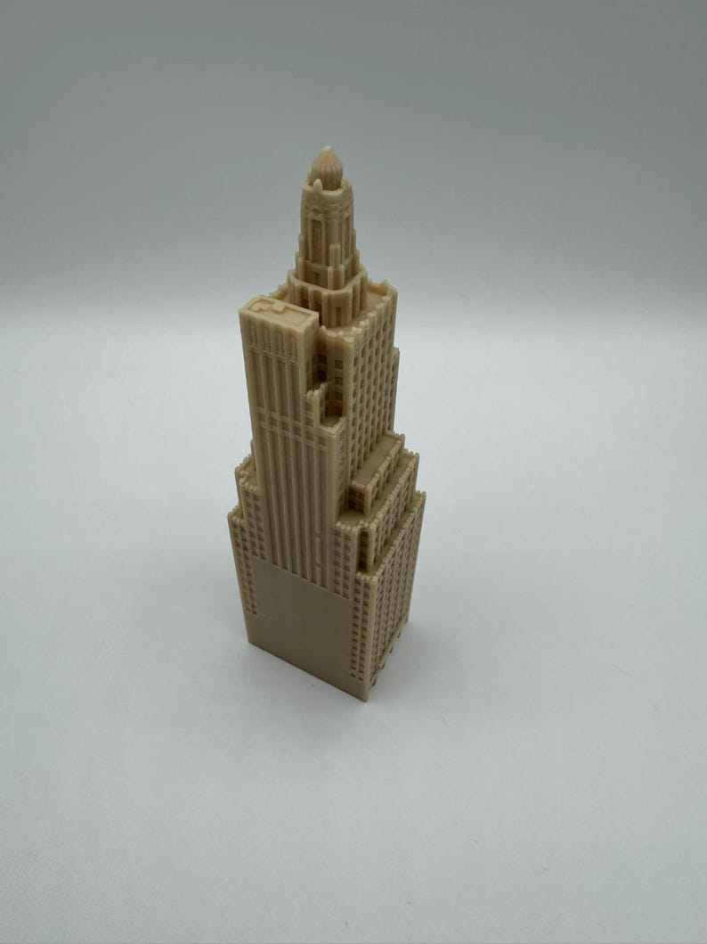 May include: A tan-colored miniature skyscraper model. The building has a detailed facade with multiple levels, windows, and architectural features. The top of the building has a decorative spire.