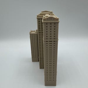 May include: Three beige miniature skyscraper models. The buildings have detailed facades with rows of windows and decorative tops. The models are arranged in a row, with the tallest one in the center and the smallest one on the left.