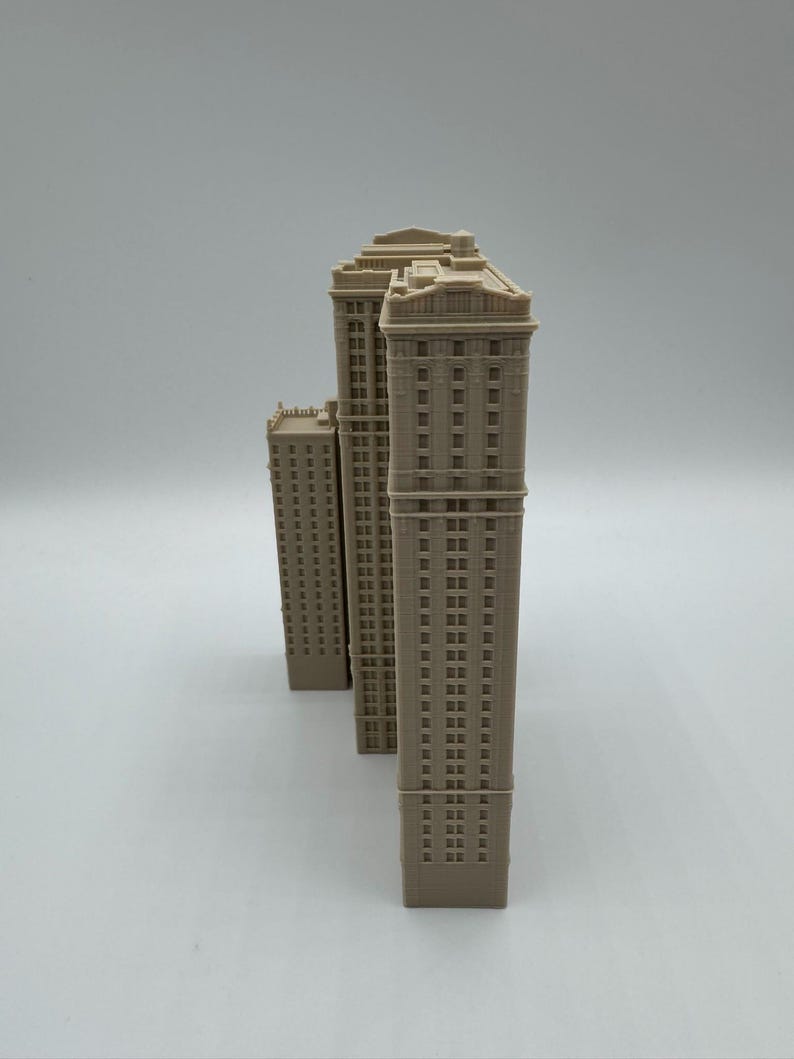 May include: Three beige miniature skyscraper models. The buildings have detailed facades with rows of windows and decorative tops. The models are arranged in a row, with the tallest one in the center and the smallest one on the left.