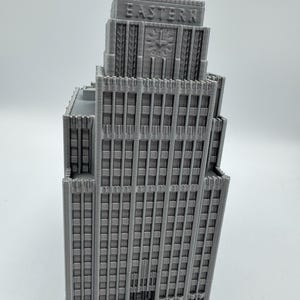 May include: A gray 3D-printed model of a skyscraper. The building features a detailed facade with numerous windows and vertical columns. The word "EASTERN" is printed on the top of the building. A clock design is visible on the building's facade.