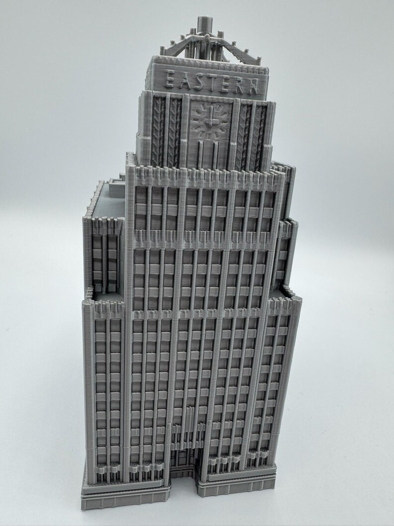 May include: A gray 3D-printed model of a skyscraper. The building features a detailed facade with numerous windows and vertical columns. The word "EASTERN" is printed on the top of the building. A clock design is visible on the building's facade.