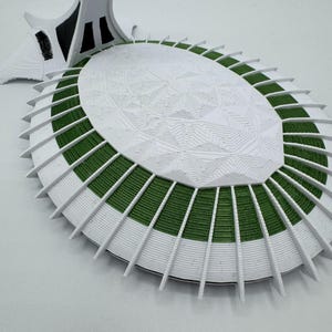 Puede incluir: Maqueta arquitect&oacute;nica blanca y verde con un dise&ntilde;o detallado y en capas. El modelo presenta una base ovalada con secciones verdes y soportes blancos radiantes. Una estructura alta y blanca se eleva desde la base, lo que se suma al tema arquitect&oacute;nico general.