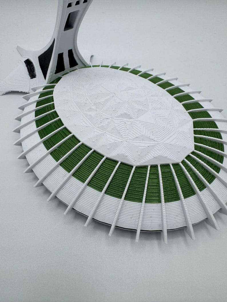 Puede incluir: Maqueta arquitect&oacute;nica blanca y verde con un dise&ntilde;o detallado y en capas. El modelo presenta una base ovalada con secciones verdes y soportes blancos radiantes. Una estructura alta y blanca se eleva desde la base, lo que se suma al tema arquitect&oacute;nico general.