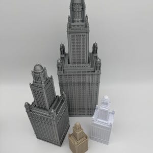 May include: A collection of miniature skyscraper models in various sizes and colors, including gray, beige, and white. The models have detailed architectural features and are arranged on a white surface. A wooden ruler is at the bottom.