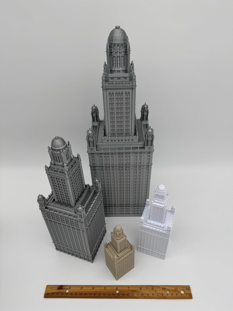 May include: A collection of miniature skyscraper models in various sizes and colors, including gray, beige, and white. The models have detailed architectural features and are arranged on a white surface. A wooden ruler is at the bottom.