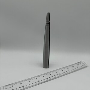 May include: A silver metal ring sizer tool with markings from 0 to 13. The tool is about 5 inches long and is lying on a white surface next to a ruler.