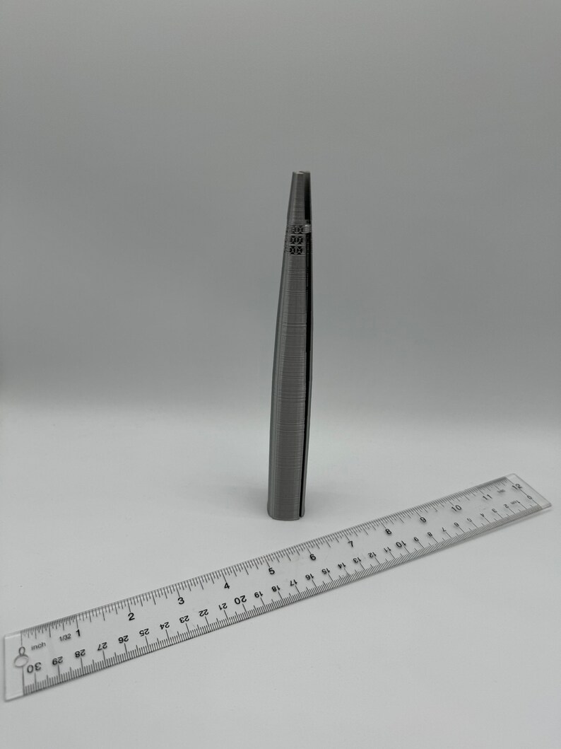 May include: A silver metal ring sizer tool with markings from 0 to 13. The tool is about 5 inches long and is lying on a white surface next to a ruler.