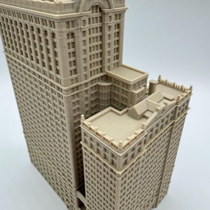 May include: A beige, detailed architectural model of a multi-story building. The model features intricate window and facade details, with a stepped design. The building has a classic, ornate style, with a flat roof.
