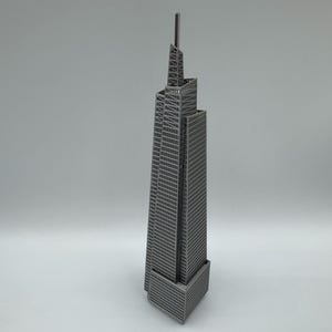 May include: A gray, miniature skyscraper model with a tall antenna. The building has a rectangular base and a detailed facade with many windows. The top section features an intricate, open-frame design.