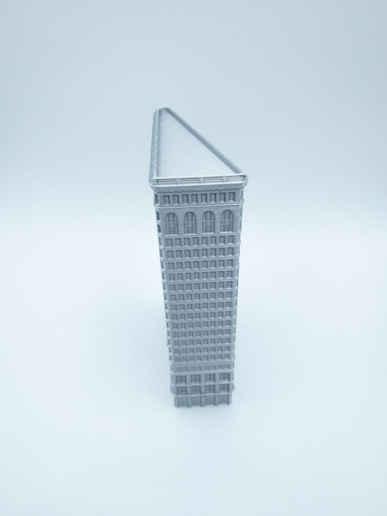 Puede incluir: Un modelo plateado del Flatiron Building en la ciudad de Nueva York. El edificio es un rascacielos triangular con una azotea plana y muchas ventanas.