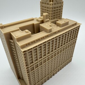 May include: A tan-colored architectural model of a building with a clock tower. The model features detailed windows, columns, and architectural elements. The clock tower has four clock faces. The building is a detailed replica.