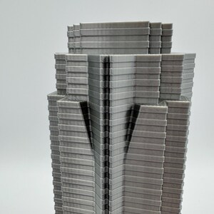 May include: A gray, layered architectural model of a skyscraper. The building has a tiered design with a series of horizontal lines creating a textured appearance. The model is set against a plain white background.